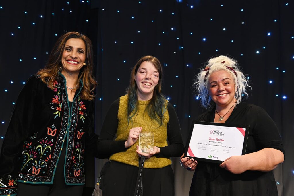 Zoe Toole from Plumpton College holding a glass trophy, flanked by two women presenting her Higher Education Student of the Year Award certificate on stage
