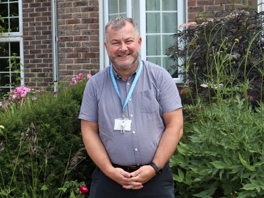 Smiling man wearing a checked shirt and staff lanyard, standing in a garden in front of a brick building