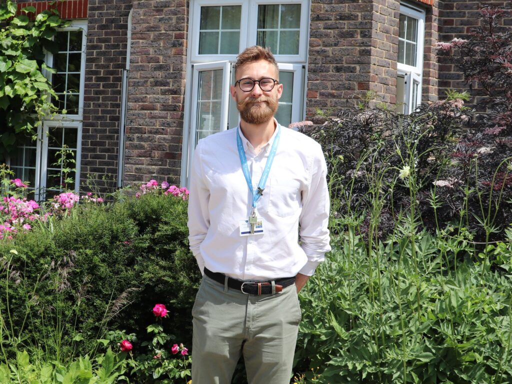 A bearded man wearing glasses and a lanyard stands in a garden in front of a brick building