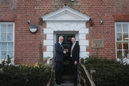 Two men in suits shake hands outside a brick building entrance marked 1766, near a Reception sign