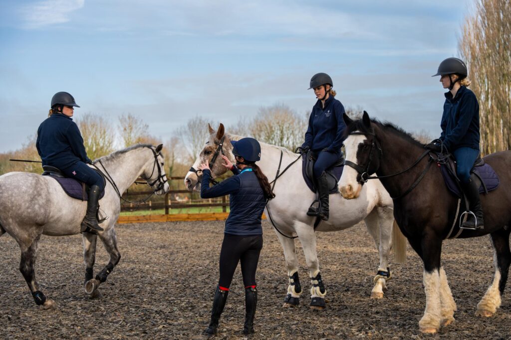 An instructor adjusts a horse's bridle as three riders in helmets sit mounted nearby in an outdoor arena